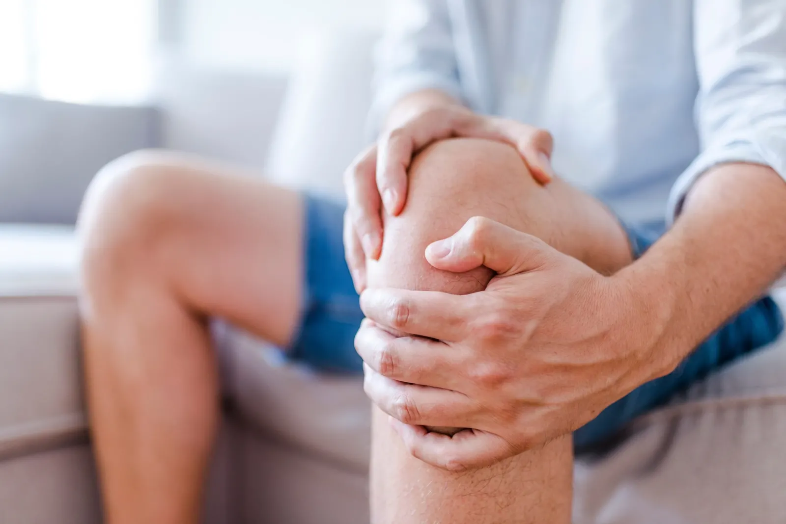 Man seated on a sofa gripping his painful knee with both hands